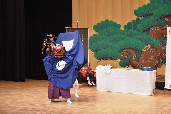 青い布をまとった胴体で赤い顔の獅子舞いがステージ上で舞っている写真
