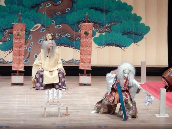 大きな松の絵が描かれた幕が見えるステージ上で右側に白髪の白い天狗のお面を被った人物が舞を舞っており、左側の着物を着た人物が座って舞の様子を見ている写真