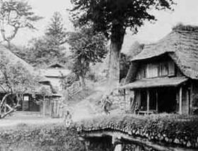 道を挟んで2つの茅葺屋根の家が建ち、手前に橋が架かっている昔の白黒写真
