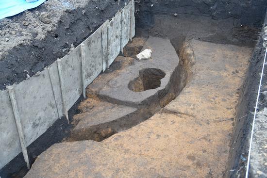 縦長に掘削したあとに、左側に弧状に大きな溝があり、内側に円形の深い穴がある発掘の写真
