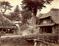 一本の大きな木の手前に茅葺屋根の家が左右に建っており、その手前に木材で造られた橋が架かっている白黒の写真