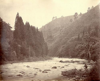 山の麓に流れている川があり、右の川辺に人がしゃがんでいる昔の風景の白黒写真