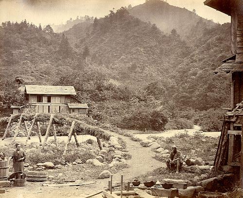 山の麓にある2階建ての民家と右手前に建つ民家の間の細い道を挟んだ左側に立っている女性と、右側に座っている男性が映っている昔の暮らしの白黒写真