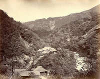 山の谷間の右側に川が流れており、左側に茅葺屋根の民家が建ち並んでいる白黒写真