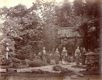 背景を樹木に覆われており、手前に池がある庭の左側に石塔や植木がある帯笑園の庭に着物を着た4名の日本髪の女性と2名の男性が立っている昔の白黒写真