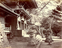 左側にある大きな寺の門の両側に、大小の灯篭があり、門の3段下に男性が座って門を見つめている白黒写真