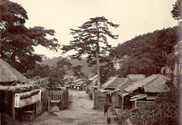 山の麓の民家の集落が通路を挟んで左右に分かれて建っており、左側はかやぶき屋根のお店が建ち、左側手前は瓦屋根と茅葺屋根の家が建ち、中央左右に門が構えている右手前に男性が立っている白黒写真