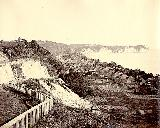 奥右側にカーブした陸地や、左側の小高い丘に柵が設置されている当時の横浜の海岸沿いを高台から撮影した白黒写真