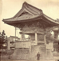 石で造られた土台に瓦屋根で覆われた大きな梵鐘の右下に、ちょんまげを結って着物を着た男性が佇んでいる白黒写真