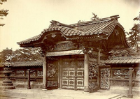 瓦屋根で中央が盛り上がっており、細かい装飾が施された門構えの屋敷の門を右斜めから撮影した白黒写真