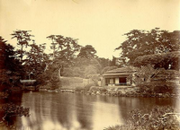 周囲に樹木や植木が植えられている庭園の手前に池があり、奥に橋、橋を渡った右側に茶室の建物がある白黒写真
