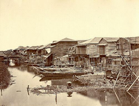 水路沿いの右側に2階建ての民家が建ち並んでおり、水路上には細長い漁船が停まって、手前の湿地に1名の男性、右側に男女が座っている白黒写真