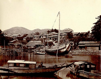 民家が建ち並ぶ手前の港に大きく反った船があり、手前の2つの漁船に男性が乗ってカメラを向いている白黒写真