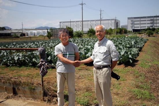 内海則行さんと作業服姿の推進委員の男性が畑の前で握手を交わしている写真