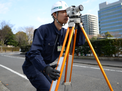 作業着とヘルメットを頭に着けた男性が道路に三脚を立てて測量の器械を覗いている写真