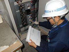 青い作業着を着てヘルメットを被った男性が、資料を見ながら電気回線のついている機械の点検をしている写真