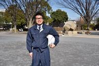 紺色の作業着を着た男性が左わきにヘルメットを抱えて立っている写真